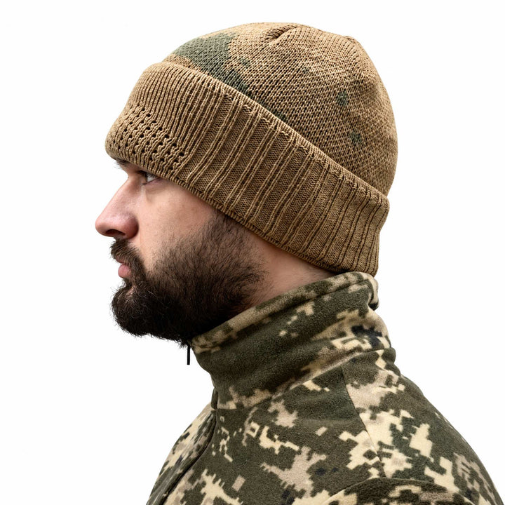 Khaki wool balaclava worn as a beanie with the lower face section folded into a wide ribbed cuff, leaving the face uncovered and showing the green camouflage knit pattern.
