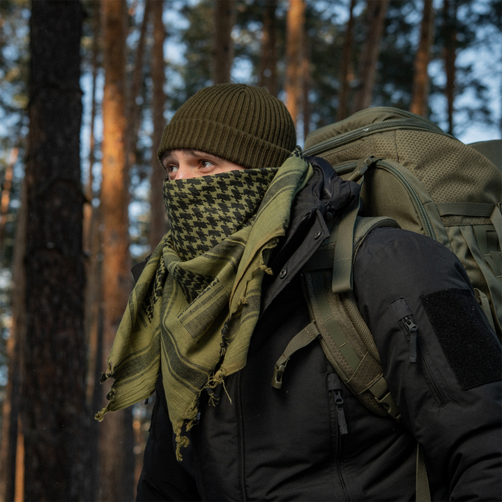 A person hiking in a forest wearing an olive green beanie, a green and black houndstooth patterned face scarf, and a large olive green backpack.