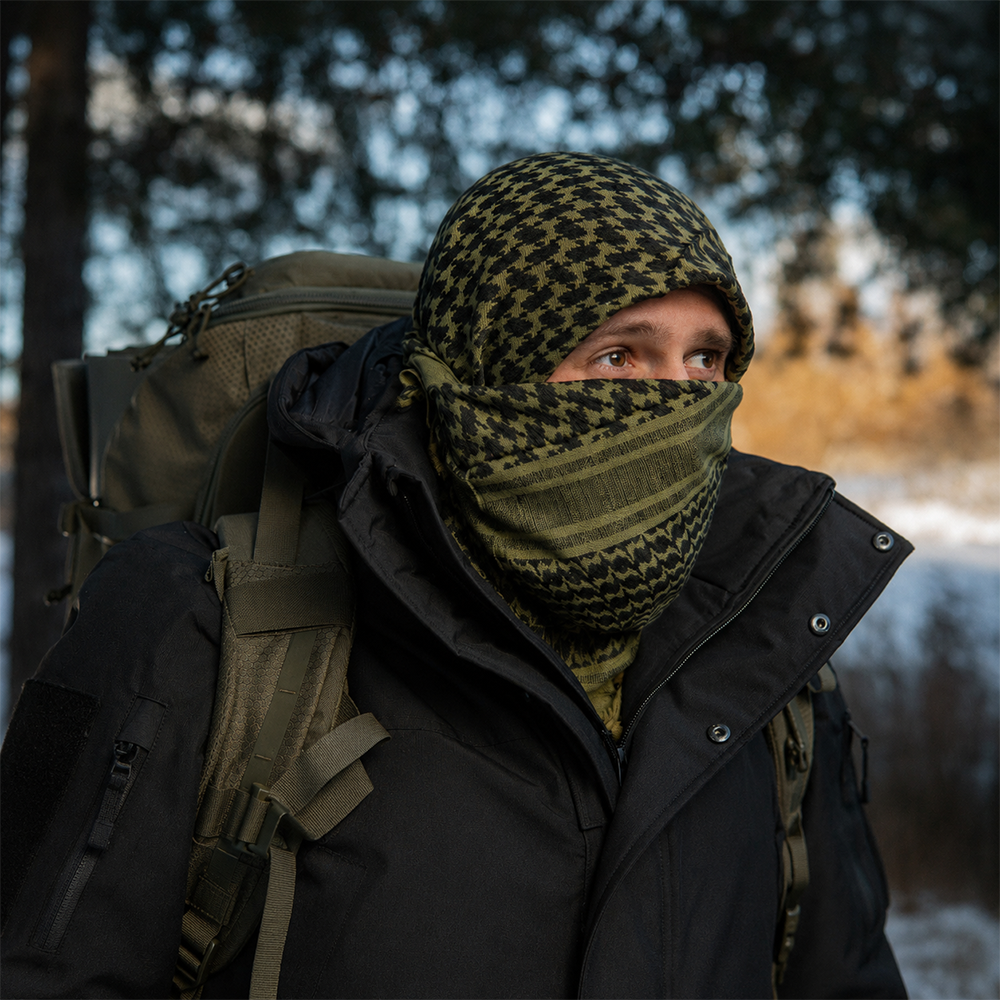 A person outdoors in a snowy forest wearing a black and olive green patterned balaclava and matching neck gaiter, a black winter jacket, and a large olive green backpack.
