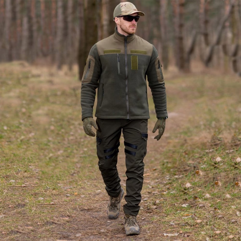 A man walking in a forest wearing a khaki tactical jacket.
