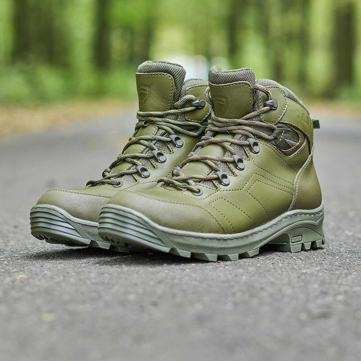 A pair of olive tactical shoes placed on a road in a forest setting, showcasing their rugged design and high-quality leather construction.