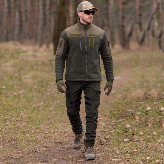 A man walking in a forest wearing a khaki tactical jacket.