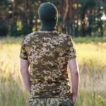Rear view of a man wearing a pixel-pattern camouflage t-shirt in a forest setting.