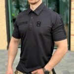 Man standing outdoors wearing a black tactical polo shirt, viewed from the front.