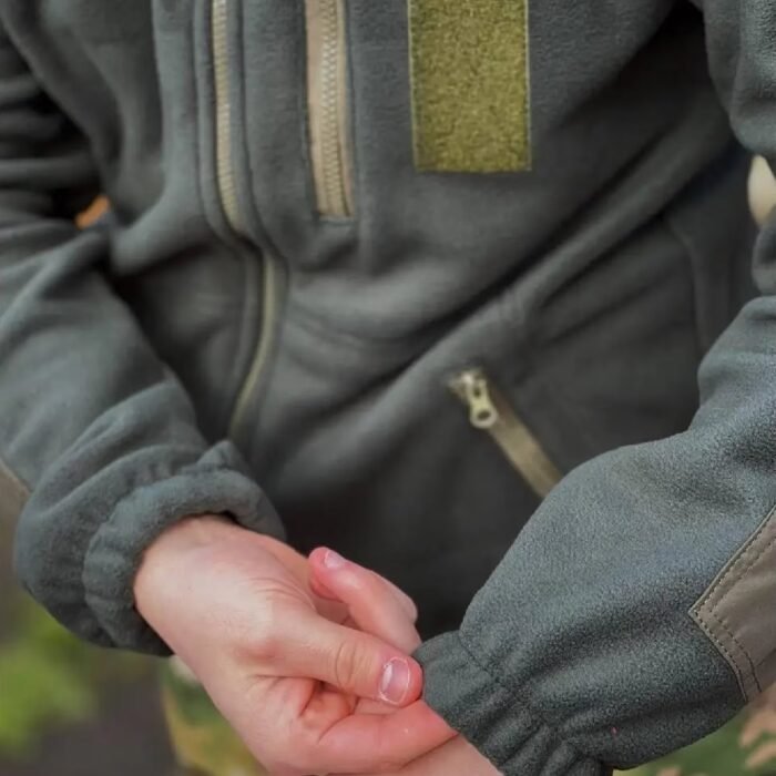 Close-up of hands with a detailed view of the zippered pocket and fabric texture on a tactical fleece jacket.