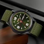 A tactical men's wristwatch with a green fabric strap and black steel case, placed on a wooden surface. The watch features a large, easy-to-read dial with day and date display.