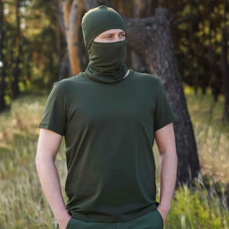 Full-body view of a man in a khaki t-shirt and balaclava standing confidently in a forest.