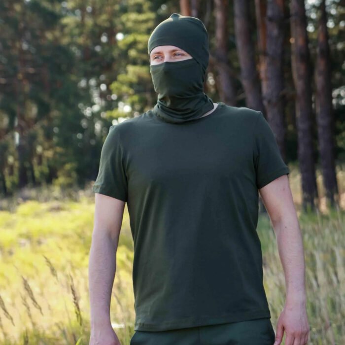 Front view of a man in a khaki t-shirt and matching balaclava standing in the woods.