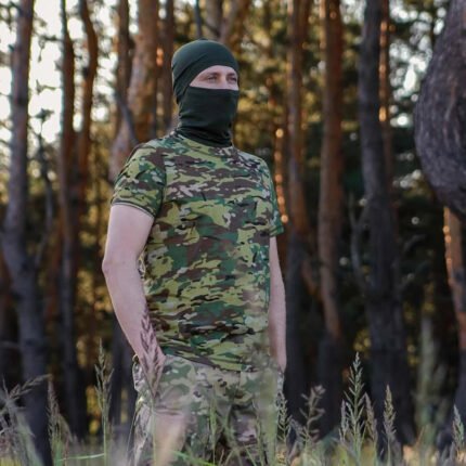 Full-body view of a man wearing a multicam t-shirt and balaclava in a forest.