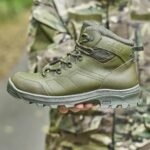 A person in camouflage holding an olive Temp tactical boot, displaying its reinforced structure and side details.