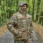 A man wearing a multicam military jacket, featuring multiple pockets, insulation, and a tactical design.